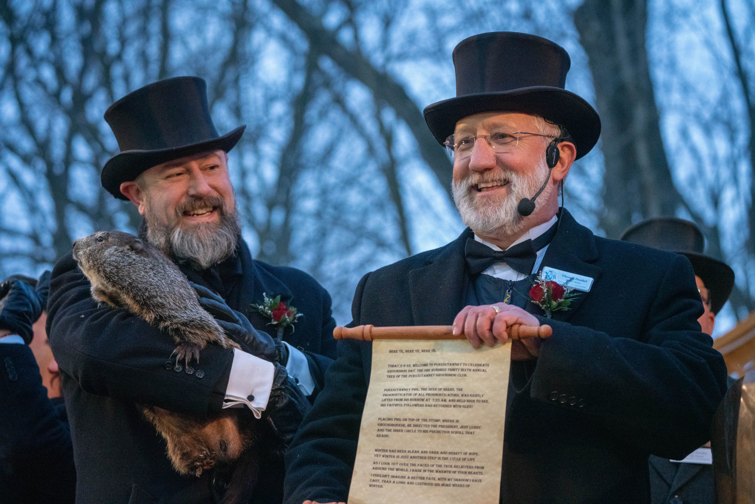 Two men wearing top hats and historical costumes. One man holds Punxsutawney Phil, the groundhog; the other holds a scroll of paper.