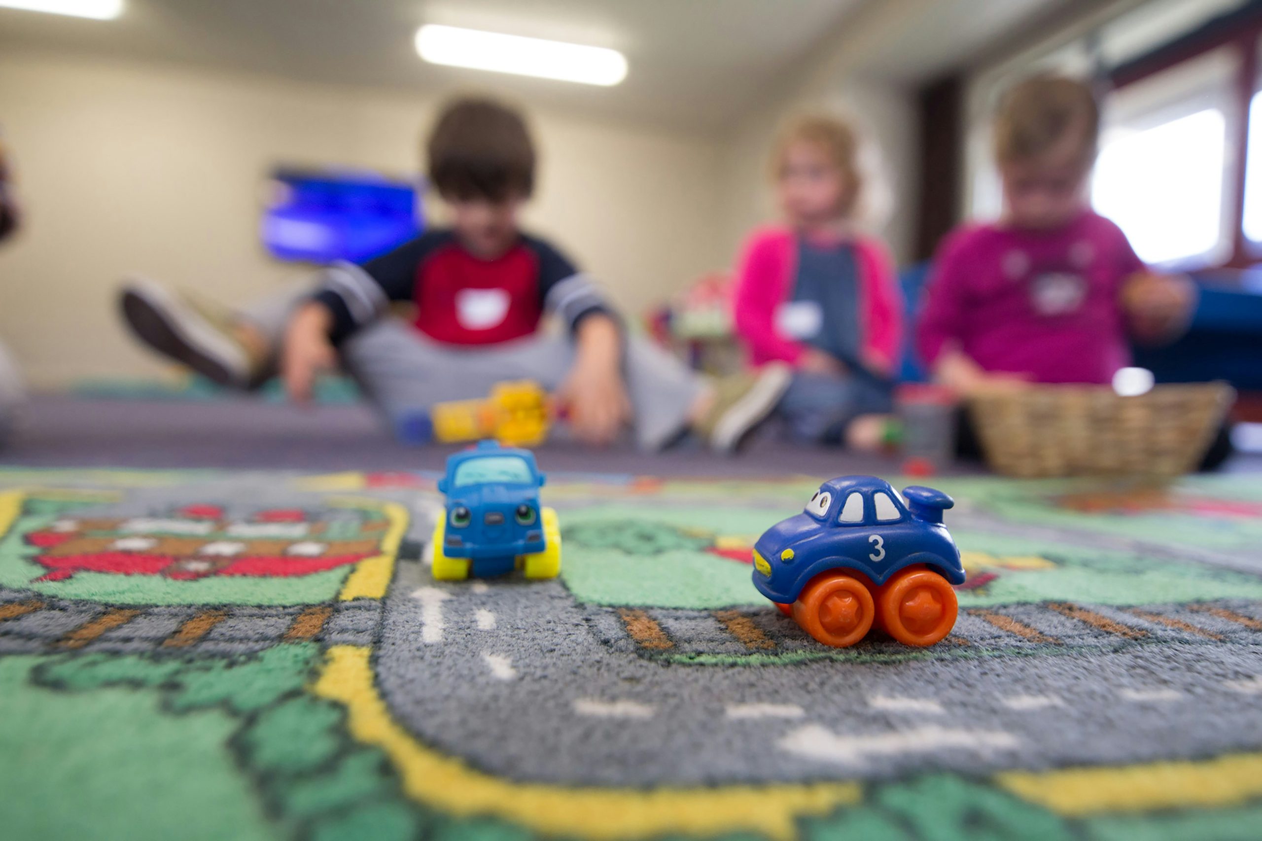 three children sit on a carpet in the background of toy cars lined up on a road/carpet