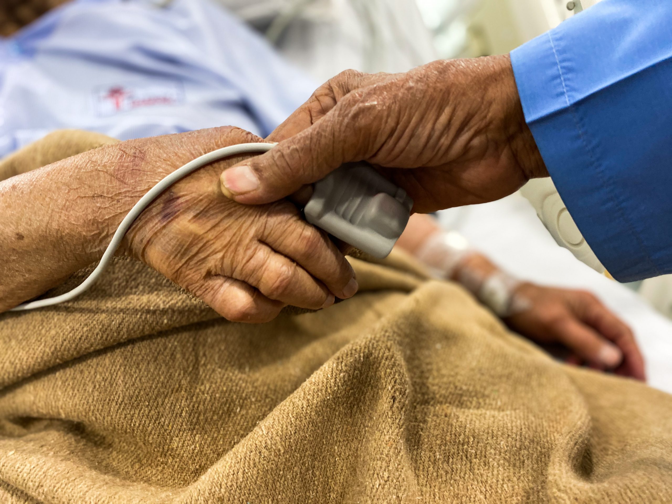 two people holding hands in hospital, with one person lying in a bed in a gown under a blanket with a monitor on their index finger