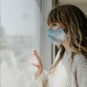 Young woman with a cloth mask on her face places her hand on window and looks outdoors.