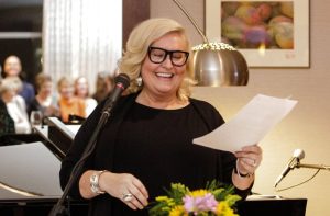 A blonde woman with dark framed glasses and sheet of paper in hand laughing at the podium with a group of people behind her