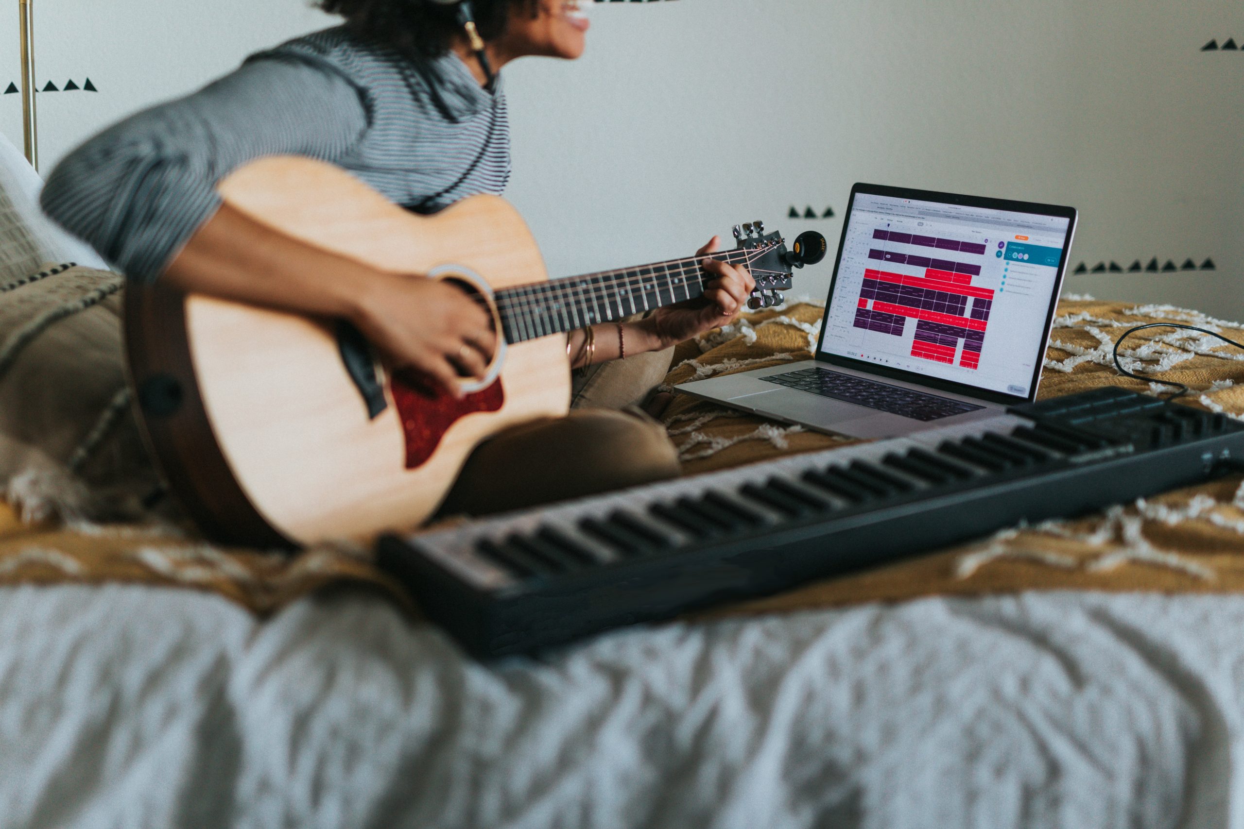 Photo of a person playing the guitar and keyboard in front of a laptop with music software open.