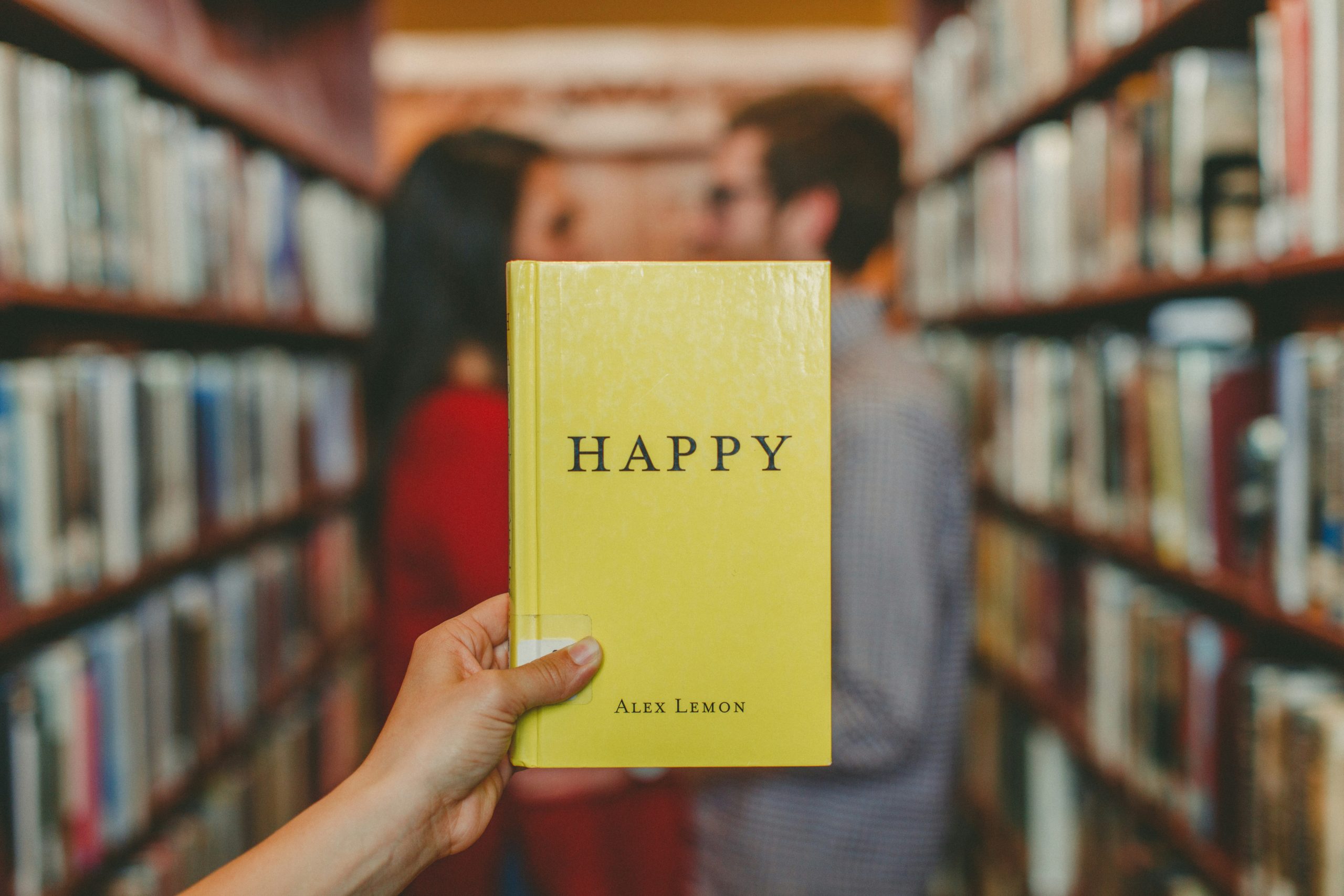 Image of a person holding up the book 'Happy' by Alex Lemon in front of two people in a library.