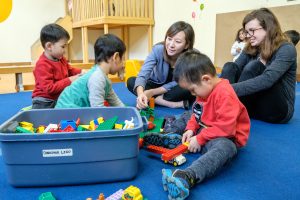 2 adult women sitting on the floor with 3 children playing with legos