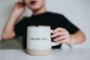 Photo of a person in a black shirt holding a white mug that reads, &quot;see the good.&quot;