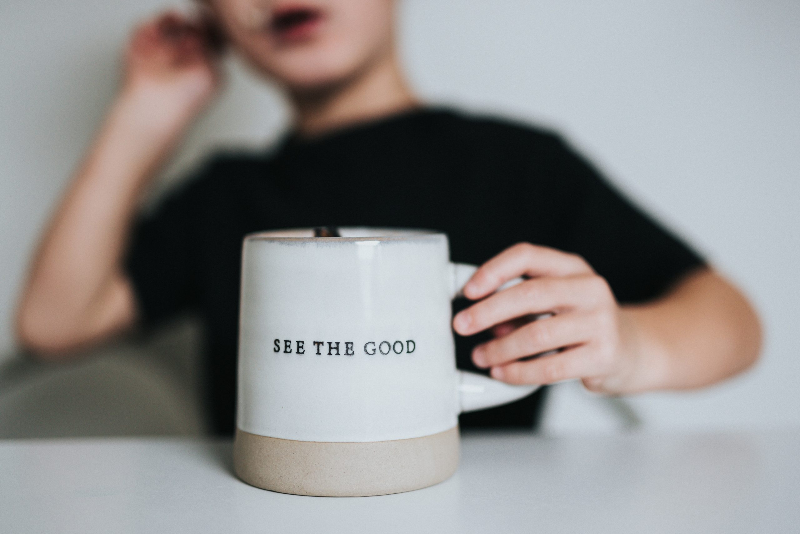 Photo of a person in a black shirt holding a white mug that reads, "see the good."