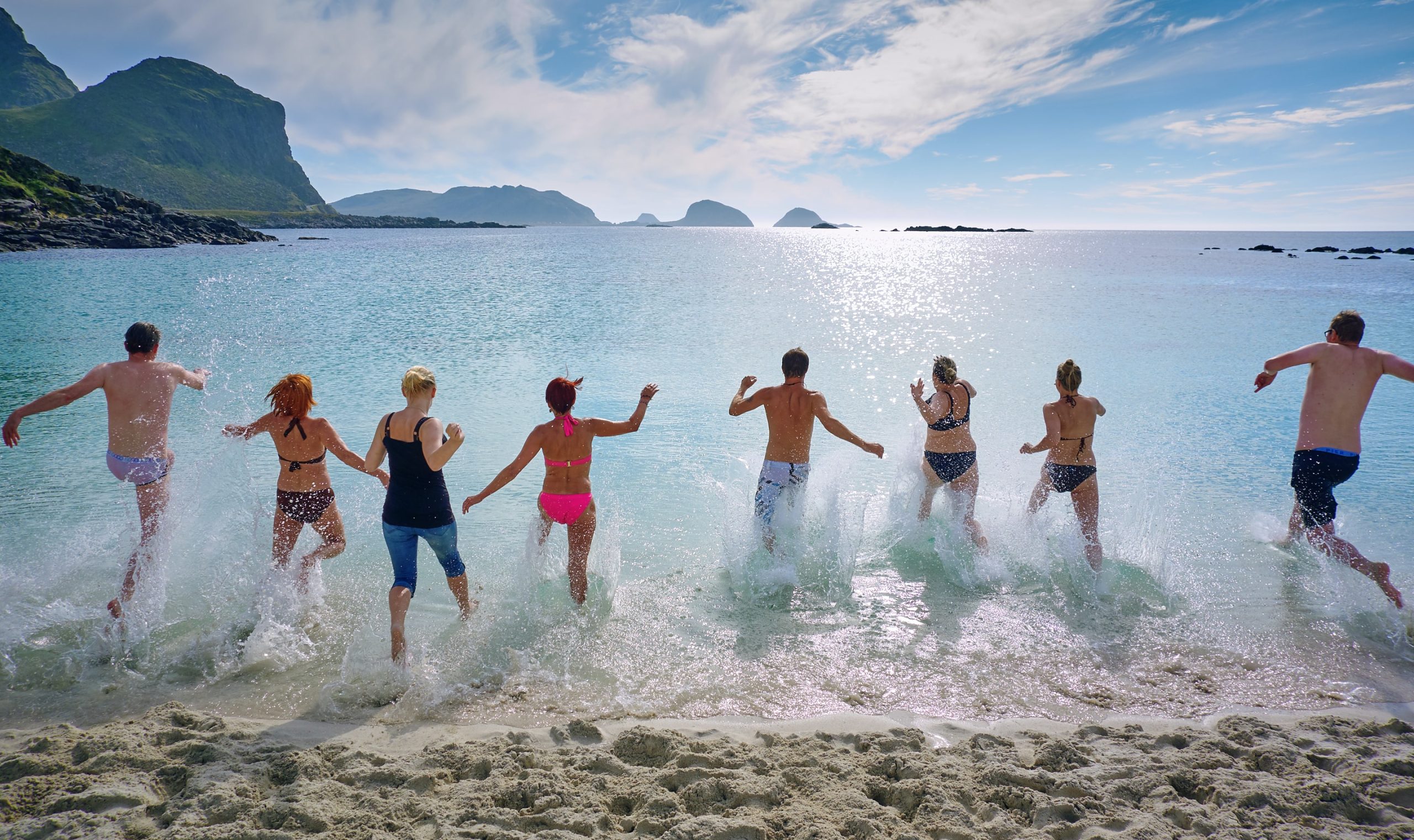 A group of adults in swimsuits at the beach, running into the water, on a sunny day with cliffs and mountains in the background.