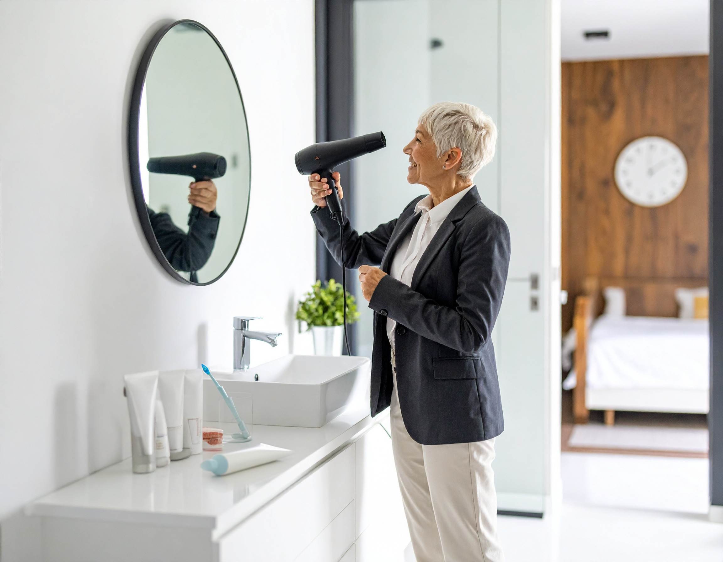 A professionally-dressed woman with short hair using a blowdryer in front of a bathroom sink and mirror, with a bedroom in the background.