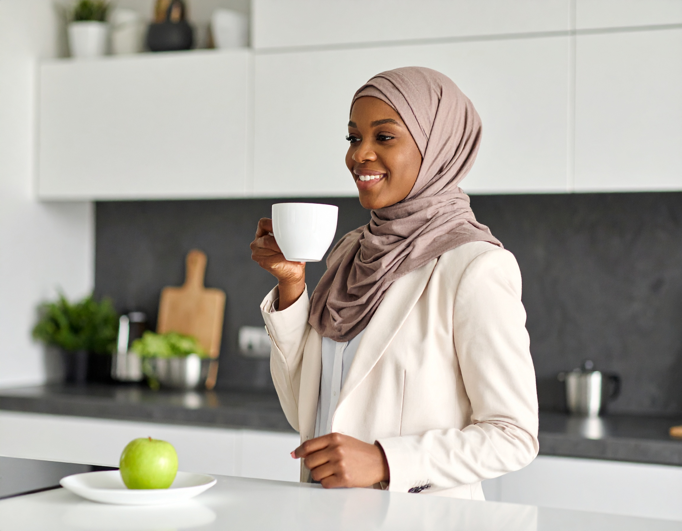 A young woman dressed in professional clothes and a hijab, drinking coffee in her kitchen, with an apple on the plate in front of her.