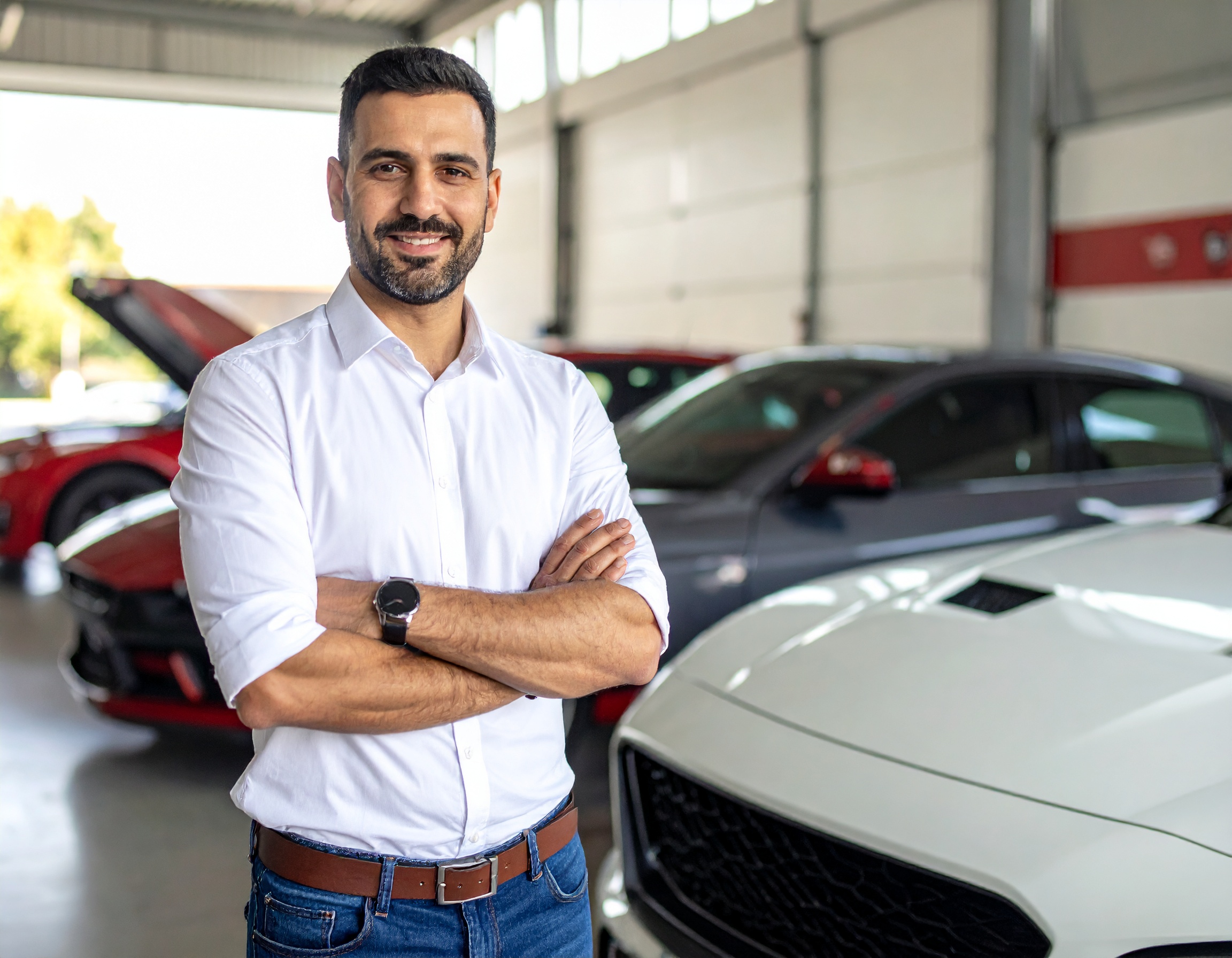 A man in his late thirties standing in a mechanic shot in front of a variety of cars.