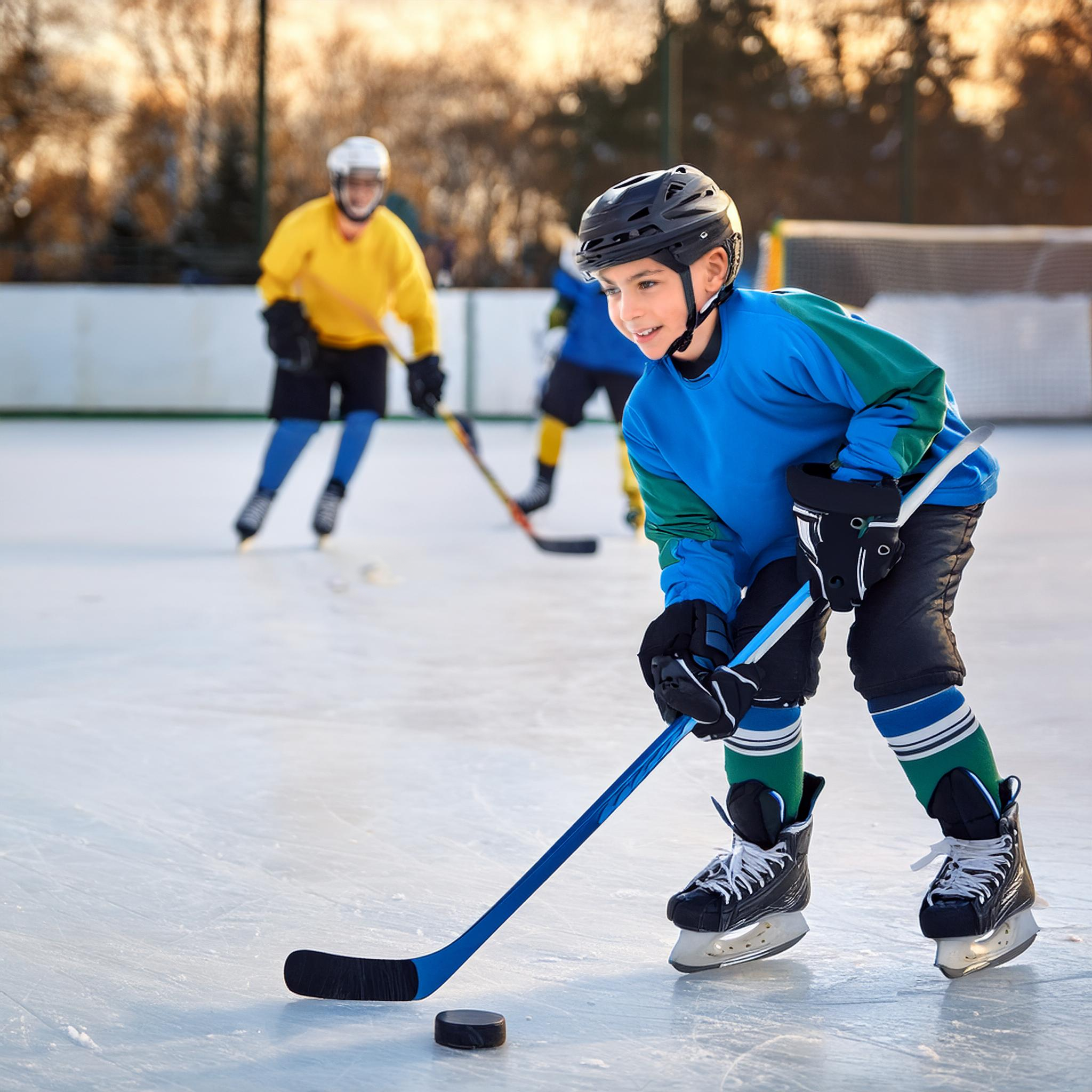 A young Hispanic boy holding a hockey stick in front of a puck on an outdoor hockey rink.