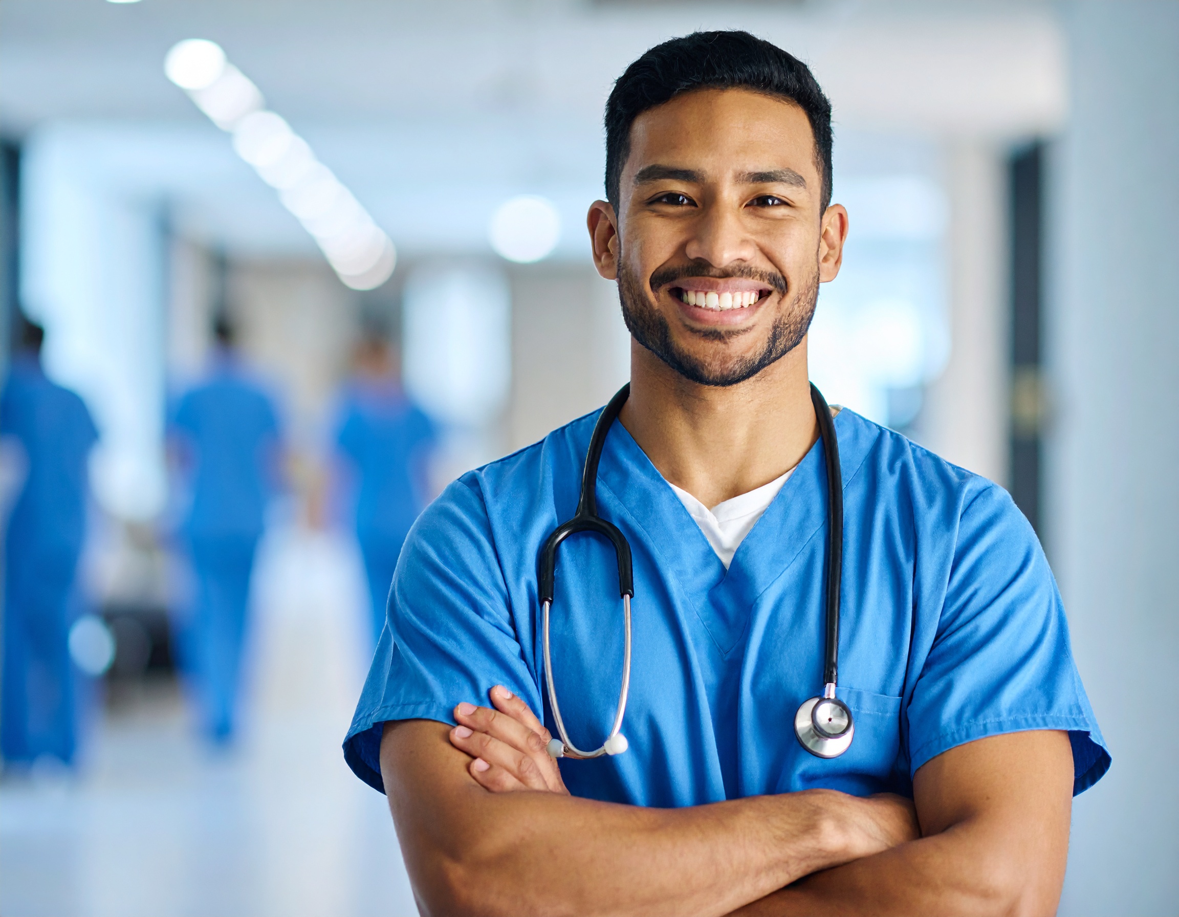 A smiling young man wearing nurse scrubs and a stethoscope in a hospital setting.