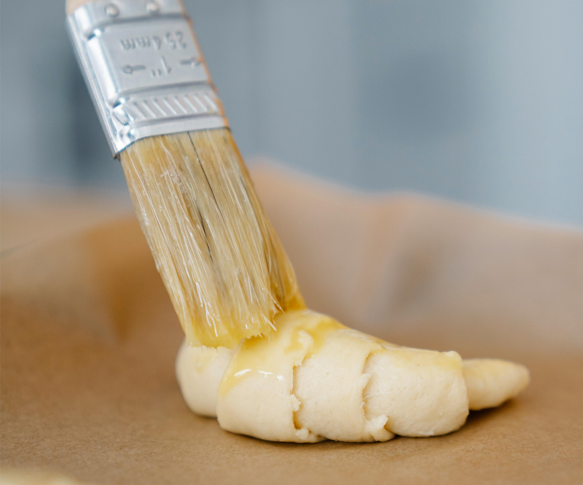 A person brushing butter onto a raw croissant.