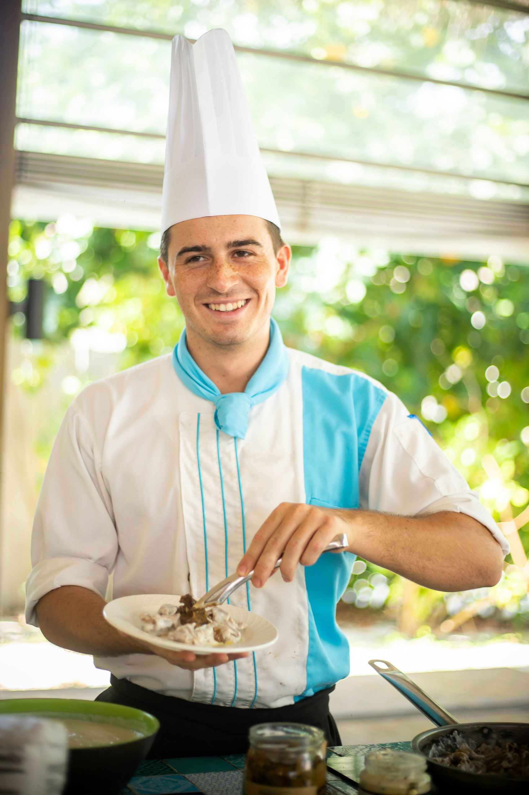 A man in chef's hat with a plate of food in a bright kitchen.