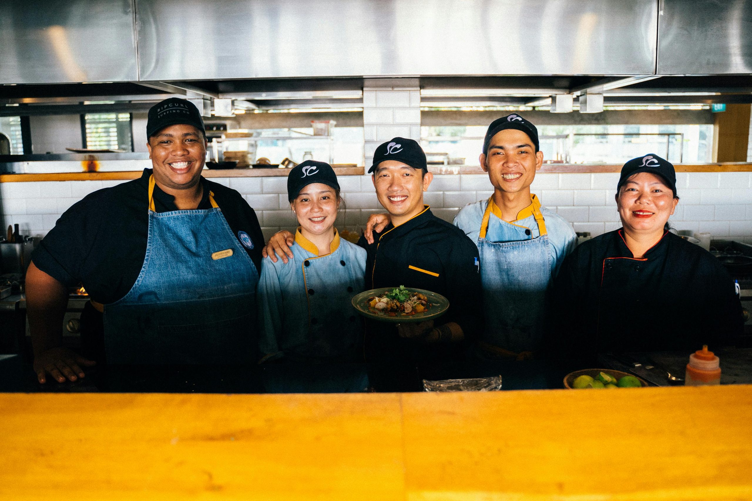 A crew of smiling restaurant workers posing with a finished dish in their kitchen.