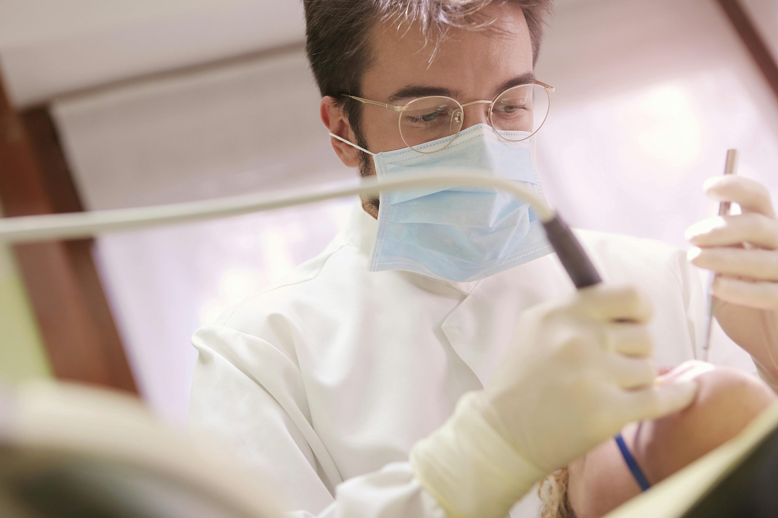 A male dentist wearing a medical mask, working with dental cleaning tools on a patient.