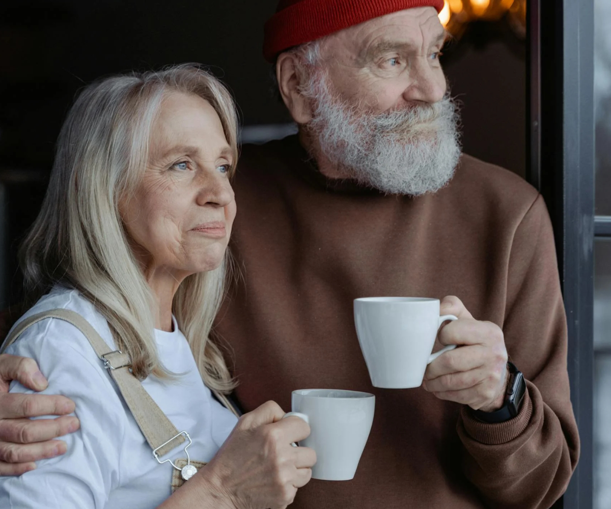 Two older adults holding coffee mugs while looking out the window.