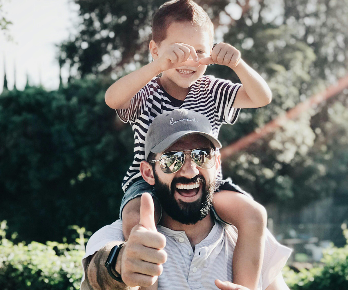 A man giving a thumbs-up while carrying his young son on his shoulders.