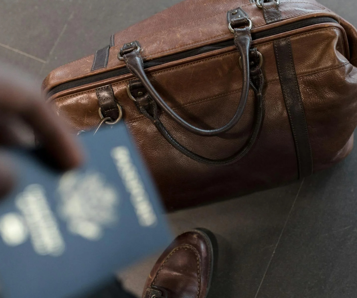 A person holding a passport over a brown duffel bag.