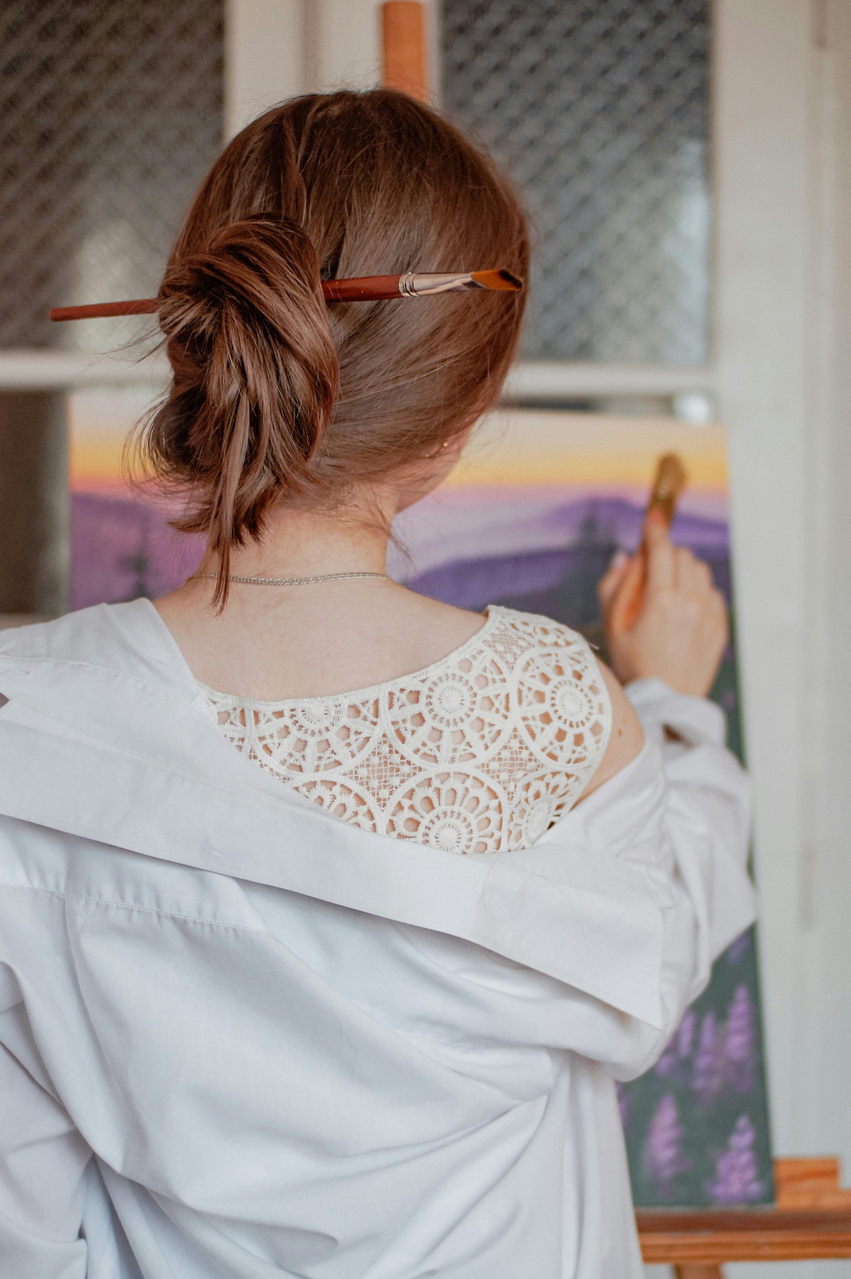 A woman with a paintbrush in her hair, painting a nature scene on a canvas.