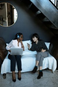 Two women sitting on couch - one using a laptop and the other texting on a cellphone