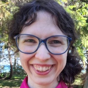 Portrait of a brown-haired woman of European descent with a light skin tone, wearing glasses and pink clothes, in front of pine trees and a lake.