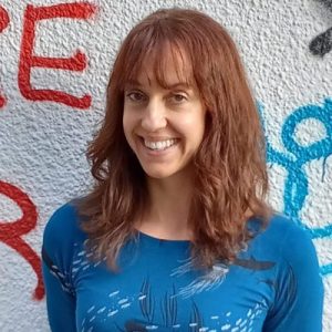 Portrait of an auburn-haired woman with a light skin tone, wearing blue clothes, in front of a wall with red and blue paint.