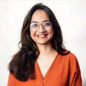 Portrait of a black-haired woman from India with a medium skin tone and glasses, wearing a rust-orange shirt.