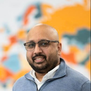 Portrait of a person with a medium-dark skin tone and a black beard, wearing glasses and grey and white clothes, in front of a multicoloured background.