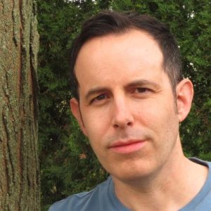 Portrait of a man with black hair and a medium-light skin tone, wearing a blue shirt, with trees in the background.
