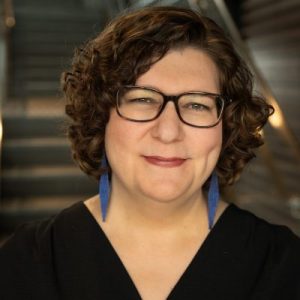 Portrait of a woman with brown hair, a light skin tone, glasses, blue earrings, and a black shirt, in front of a staircase.