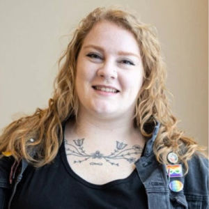 Portrait of a Métis person with light skin, red hair, and traditional floral chest tattoo, wearing a jean jacket with a rainbow pin and black shirt.