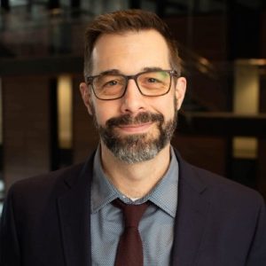 Portrait of a brown-haired man with a beard and a light skin tone, wearing glasses and a suit, in front of a staircase in a building with brown bricks.