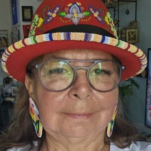 Portrait of an Anishinaabe (Ojibwe) and Ongweh’onweh (Oneida) woman with a brown skin tone and brown hair, wearing a red hat with colourful beadwork, beaded earrings, and glasses.