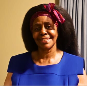 Portrait of a black-haired woman with a medium-dark skin tone, wearing blue clothes and a pink and purple headband, in front of a light brown background with a blue curtain.