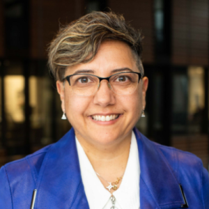 Portrait of a woman with brown hair and a medium skin tone, wearing glasses and a blue jacket with a white shirt, in a building with brown wooden panels.