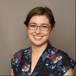 Portrait of a brown-haired woman with a light skin tone, wearing glasses and black clothes with multicoloured flowers.