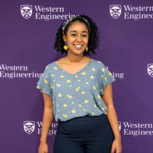 A woman with black hair and a medium-dark skin tone, wearing a black and white headband, blue and yellow clothes, and earrings, standing in front of a purple background with the words “Western Engineering.”