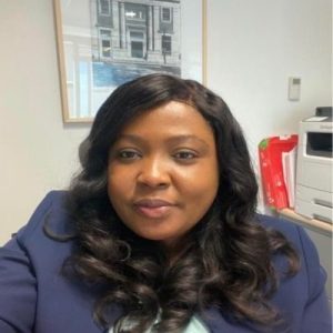 Portrait of a black-haired woman with a medium-dark skin tone, wearing a blue suit, in a white office with a black and white photo of a building.
