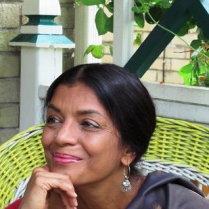 Portrait of a black-haired Indian-Canadian woman, with a medium-dark skin tone, wearing earrings and a black sari, seated outdoors.