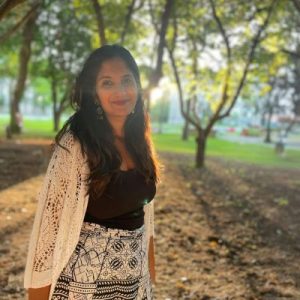 A black-haired woman from India with a medium skin tone, wearing black and white clothes and earrings, standing on a campus pathway with trees in the background.