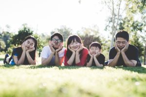 A modern family photo depicting two parents and three children from East Asia, posing on the group with their hands framing the bottom of their faces. All of their eyes are closed and some are making funny faces.