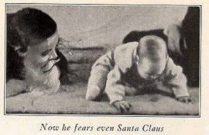 An old newspaper clipping shows a young, 18 month old boy, sitting down and leaning forward as to crawl away. An older man to the left, with a long white beard like Santa Claus, leans toward him in a friendly manner. The headline reads “Now he fears even Santa Claus”