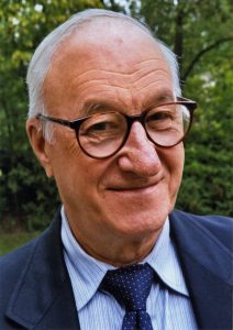 A coloured picture of a happy white man, Albert Bandura, wearing glasses with a blue suit and tie, smiling at the camera
