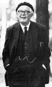 A black and white photo of Jean Piaget, an older white man wearing a three-piece suit, glasses, and a beret. He is looking upward with a look of wonderment, and a slight smile.
