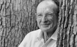 A modern black and white photo of Urie Bronfenbrenner, leaning against a tree, wearing a white collared shirt, glasses, and smiling.