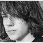 Black and white image of a melancholy teenage boy with shoulder-length wavy hair covering one eye.