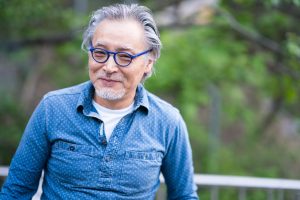 With a blurry background of green foliage, an older man smiles slightly toward the camera wearing a blue button-up shit and small blue glasses. He has a grey goatee and grey hair lightly combed back.