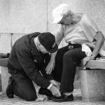 This black and white image depicts an older man kneeling down to help his wife fix her shoe. She is gently placing a hand on his arm.