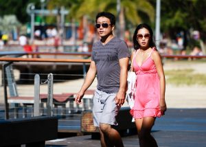 With a blurry background of a park and playground equipment, a young man and young woman hold hands as they walk down a path.